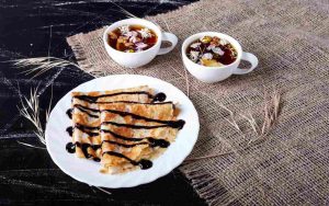 Crepes con sciroppo al cioccolato su un piatto accanto a due tazze di thè